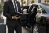 Un chauffeur consulte un dossier de formation et une carte vierge près d’une berline générique.