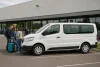 Un minibus blanc générique devant un bâtiment commercial, avec des voyageurs et leurs bagages.